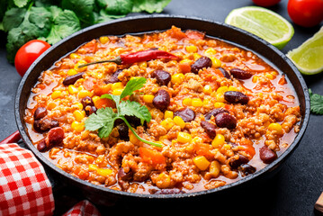 Chili con carne with beef, red beans, paprika, sweet corn and hot peppers in tomato sauce, spicy tex-mex dish in frying pan, black table background, top view