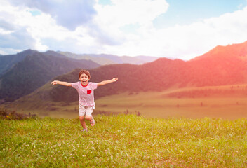 Naklejka na ściany i meble Happy little boy running and jumping on green meadow with mountain background
