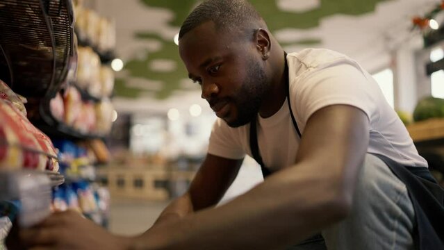 Close-up shot of a Black-skinned man in a white t-shirt and black apron crouching near a shop window and laying out goods in a large supermarket