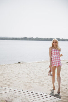 Young blonde girl look like a Barbie doll in pink mini dress walking barefoot on beach.