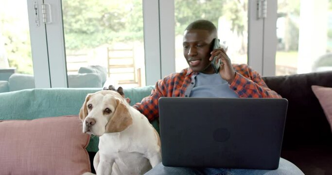 Happy African American Man On Smartphone And Using Laptop At Home, With His Pet Dog, Slow Motion