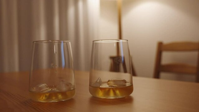 The man and woman place their glasses of whiskey next to each other on a wooden table. The scene is illuminated by dim and warm lighting in a white room.