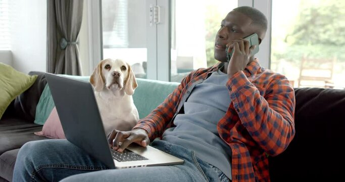 African american man talking on smartphone and using laptop at home, with his pet dog, slow motion