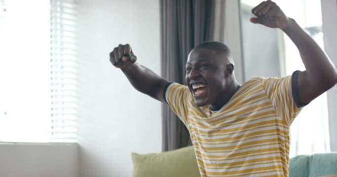 Happy african american man watching match and celebrating at home, slow motion