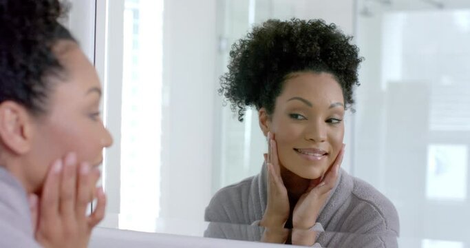 Happy biracial woman in bathrobe looking in mirror in bathroom, slow motion