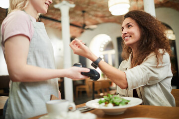 Cheerful excited young lady with curly hair sitting at table and paying for dinner with smartwatch while smiling at friendly waitress in restaurant