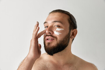 Beauty closeup shoot of enjoyed young man applying facial cream isolated over gray background.