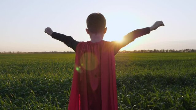 A happy child in a red cape stands against the sky during sunset. The superhero boy stands with his arms outstretched, depicting strength and success, freedom. The concept of a kid dream.