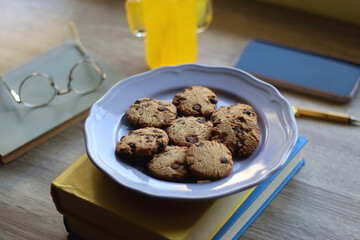 Plate of chocolate chip cookies, stack of books, reading glasses, orange soda, mobile phone and pen on the table. Hygge at home. Pastel colors, selective focus.