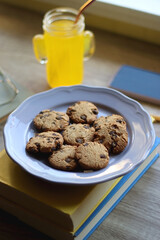 Plate of chocolate chip cookies, stack of books, reading glasses, orange soda, mobile phone and pen on the table. Hygge at home. Pastel colors, selective focus.