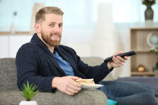 man changing tv channel with remote control while eating sandwich