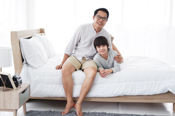 Fototapeta na wymiar Smiling Asian cute little boy is relaxing and hugging with father in bedroom at home. Family love and child educational for homeschool on holiday concept.