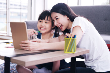 Smiling Asian mother and little girl child is drawing and Painting with wooden colored pencils on paper together in living room. She online learning with laptop. Homeschool and educational concept