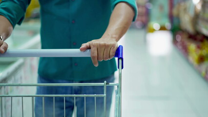 Close-up of consumer hand pushing shopping cart walking in grocery store aisle, depicting...