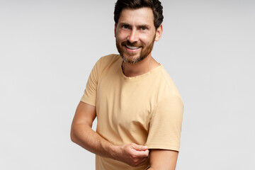 Fototapeta premium Happy bearded man model wearing stylish t shirt looking at camera and showing biceps isolated on grey background. Confident modern hipster with stylish hair smiling. Mockup 