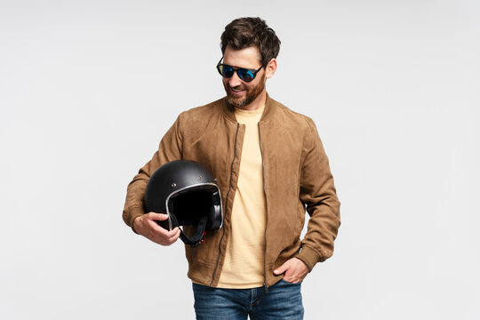 Handsome smiling bearded man, biker holding motorcycle helmet isolated on gray background. Fashion model wearing brown leather jacket, stylish sunglasses posing for pictures 