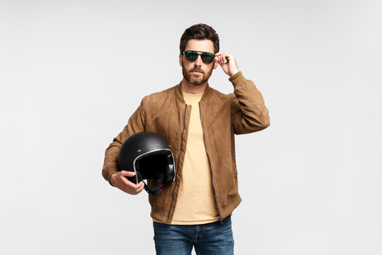 Confident serious bearded man biker holding motorcycle helmet and looking at camera isolated on gray background. Fashion model wearing brown leather jacket, stylish sunglasses posing for pictures 