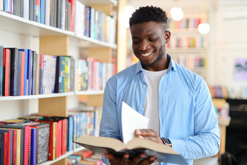 Black man in a library or bookstore holding an open book and reading it while wearing a blue...