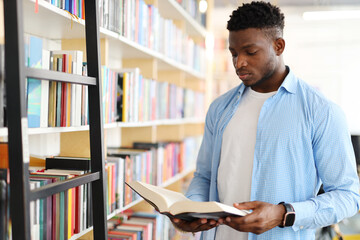 Black man in a library holding a book in their hands while wearing a blue shirt. The person is...