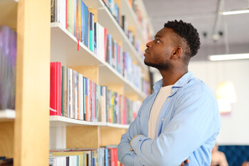 Fototapeta premium Black man standing in front of a bookshelf and looking at books