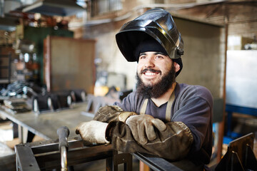 Cheerful dreamy bearded young man with dirty sleeves wearing welding mask and looking with burning...