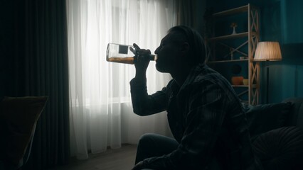 Side view of the dark silhouette of a man emptying a bottle of alcohol. A man sits in a dark room...