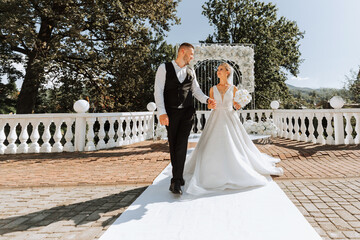 Stylish groom in white shirt and black vest and cute blonde bride in white dress in park near elegant white rose arch. Wedding portrait of newlyweds.