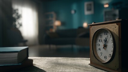 A vintage clock stands on a table close up, against a blurred background of a room illuminated by...