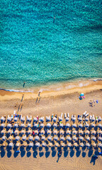 Aerial view of sandy beach with colorful umbrellas, swimming people in sea bay with transparent blue water at sunset in summer. Aerial top view on the beach, umbrellas, sand and sea waves.