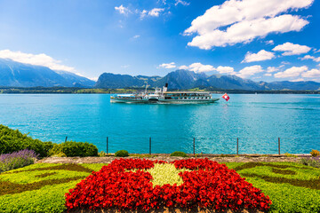 Flowerbed of the Swiss flag with boat cruise on the Thun lake and Alps mountains, Oberhofen, Switzerland. Swiss flag made of flowers and passenger cruise boat, Lake Thun, Switzerland.