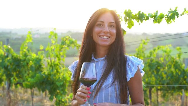 Young woman inside wineyard holding glass of red vine during sunset time and smiling on camera

