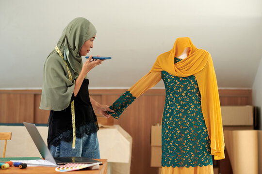 Muslim fashion designer looking at dress on mannequin and recording voice message for client