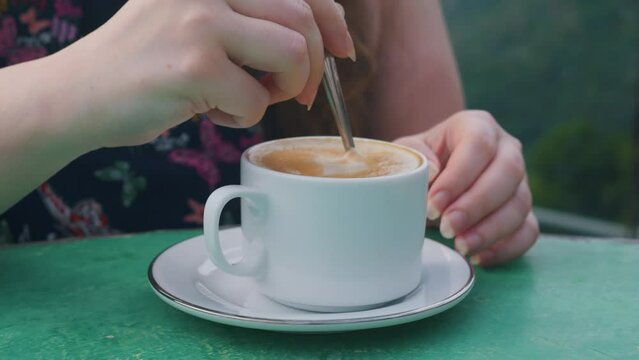 Woman hand stiring cappuccino coffee in a cup with teaspoon.