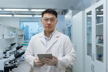 Naklejka na ściany i meble Portrait of a young Asian male scientist standing in a laboratory wearing protective glasses and gloves. He was holding a folder with documents in his hands. He looks seriously into the camera.