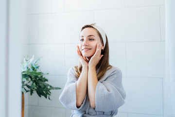 Young smiling woman applying cream mask and doing manual self massage of face in the bathroom....