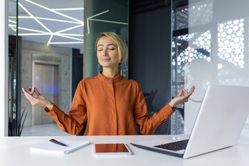 Businesswoman resting at workplace meditating in lotus position sitting at the table inside the...