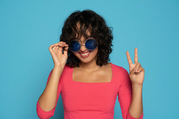 Young exotic smiling brunette woman in pink blouse posing with a sunglasses on a blue background