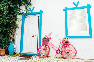 Bright pink bicycle decorated with artificial flowers standing near a white wall with closed window...
