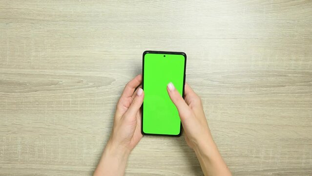 Woman using smart phone with green screen on wooden table background. Female hands scrolling pages, zooming, tapping on touch screen. top view. Office desk background. Chroma key.