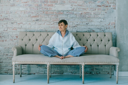 Athletic elderly woman wearing white suit sitting on sofa in lotus position
