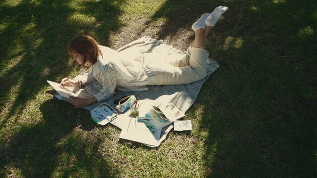 An adult inspired woman lying on the blanket in the green park and drawing in her sketchbook