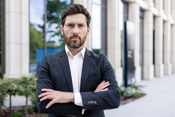 Portrait of a serious young man, a businessman, a lawyer standing in a business suit outside an office center, looking at the camera with his arms crossed. Close-up photo