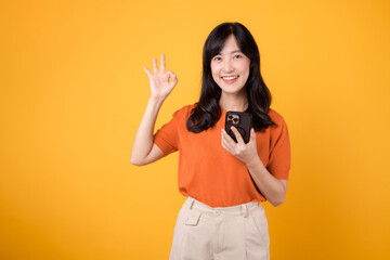 Radiate happiness with a young Asian woman in her 30s, dressed in an orange shirt, showing okay hand sign while using smartphone on yellow background.