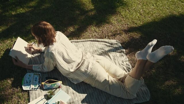 An adult relaxed woman lying on the blanket in the park and drawing in her sketchbook