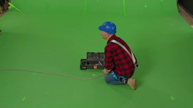 Rear view of a male builder in professional ammunition and a construction helmet sitting and repairing something on a green background