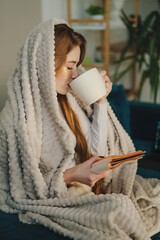 Beautiful sweet woman holding cup drinking hot coffee and reading a book at home sitting in living room on sofa enjoying holiday vacation. Relax, leisure or