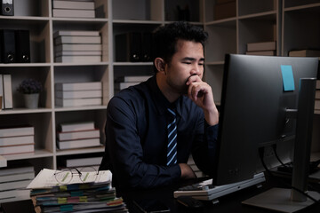 Tired young Asian businessman working overtime in the office room at night, trying to get things done before the deadline.