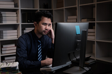 Tired young Asian businessman working overtime in the office room at night, trying to get things done before the deadline.