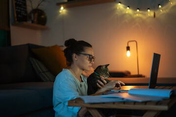 Young female freelancer student working at home at night using laptop with gray cat companion in...