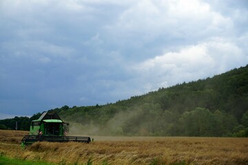 Fototapeta na wymiar Weizenernte mit grünem Mähdrescher im Einsatz beim Ernten auf Feld vor Wald und Himmel bei Sonne am Abend im Sommer